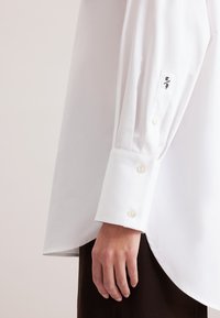 Camicia bianca a maniche lunghe con polsino a tre bottoni e cuciture decorative sottili sulla manica. Tessuto liscio e leggero.