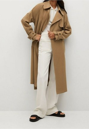 Kobieta w długim płaszczu trencz w kolorze wielbłądzim, białym prążkowanym topie, szerokich białych spodniach i czarnych sandałach typu slide, stojąca na jednolitym tle.
