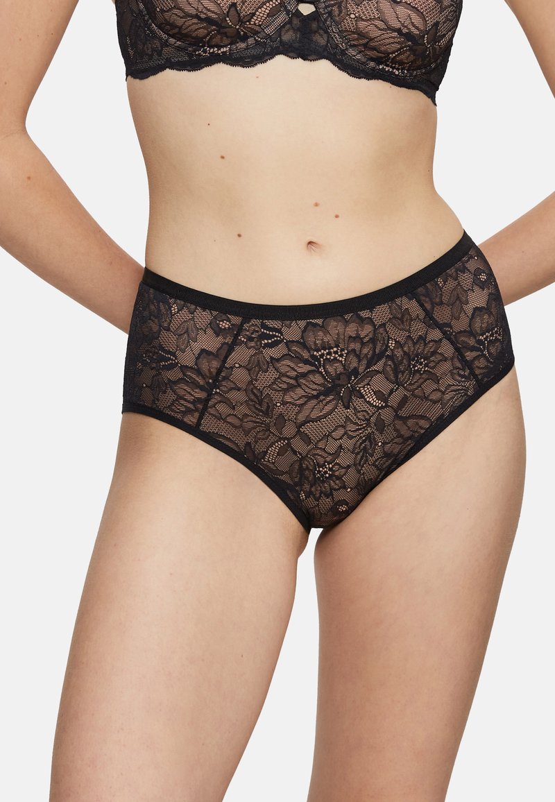 Femme portant un soutien-gorge en dentelle noire à fleurs et une culotte haute assortie en dentelle, debout avec les mains derrière le dos, devant un fond uni.