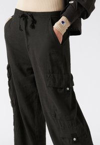 Zwarte cargo broek met een relaxte pasvorm, trekkoord taille, zijzakken en drukknoopdetails. Zachte stof met een licht gestructureerde afwerking.