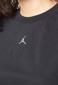 Gros plan d'une personne portant un t-shirt noir avec un petit logo blanc de joueur de basket-ball sur la poitrine, montrant le cou et les cheveux bouclés.