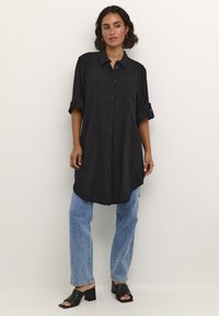 Chemise noire à boutons avec un ourlet arrondi, manches retroussées et une poche avant, associée à un jean droit bleu clair et des sandales à talons carrés noires.
