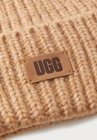 Chapeau en tricot beige avec texture côtelée et une étiquette en cuir brun cousue portant le mot "UGG" en gras. Matériau durable et doux.