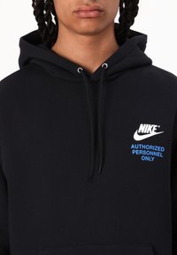 Person som bär en svart Nike-hoodie med vitt logo och blå text "Endast auktoriserad personal" på bröstet, vänd framåt.