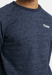 Man som bär en marinblå spräcklig sweatshirt med "Originals" broderat på höger bröst, med rund halsringning och raglanärmar.