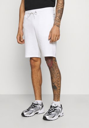 Pantalones cortos de algodón blancos con una cinturilla elástica y cordón, combinados con zapatillas deportivas negras y plateadas. Tatuajes notables en la pierna.
