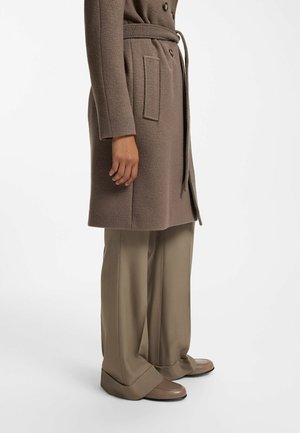 Persoon draagt een lange taupe kleurige jas met riem, beige broek met wijde pijpen en taupe loafers, staand tegen een effen witte achtergrond.
