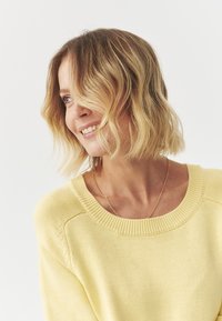 Pulover galben deschis, cu textură tricotată, guler rotund, cu margini cu nervuri. Modelul are părul ondulat până la umeri și poartă un colier simplu.