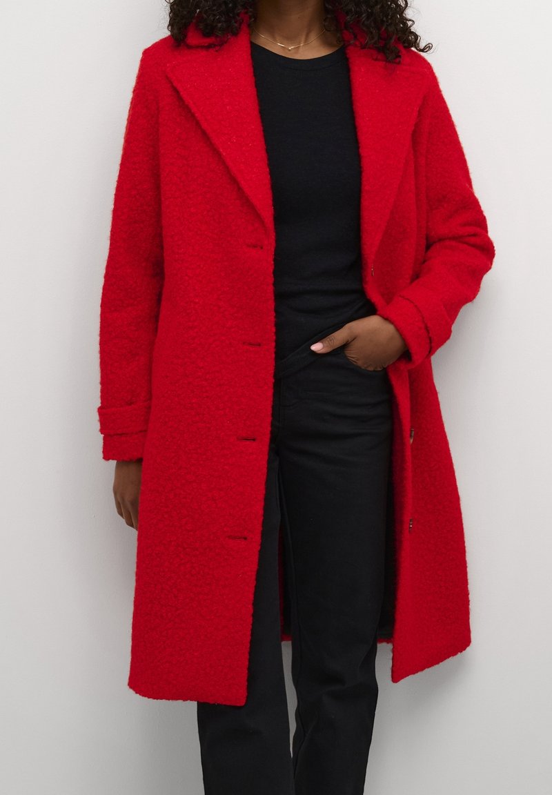 Femme portant un manteau texturé rouge vif par-dessus une chemise noire et un pantalon noir, avec une main dans la poche de son pantalon, devant un fond uni.