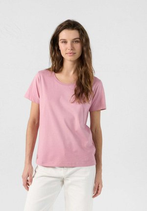 Mujer con cabello castaño ondulado, vistiendo una camiseta de mangas cortas rosa lisa y pantalones blancos, de pie contra un fondo ligero liso.