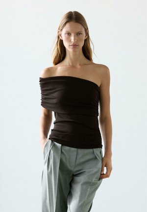 Frau mit langen Haaren, die ein schwarzes einseitiges Oberteil und hellgraue plissierte Hosen trägt, steht mit den Händen in den Taschen vor einem einfarbigen Hintergrund.