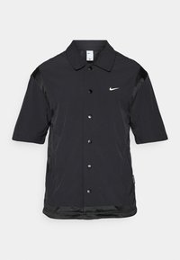 Fekete, rövid ujjú dzseki gombos elülső résszel, hálós oldalsó panelek és egy kis fehér Nike logó a bal mellrészen. Simább textúra.