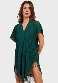Cubre-vestido estilo kimono verde con cuello en V, mangas cortas, patrón floral y dobladillo con flecos. Tejido ligero, diseño abierto.