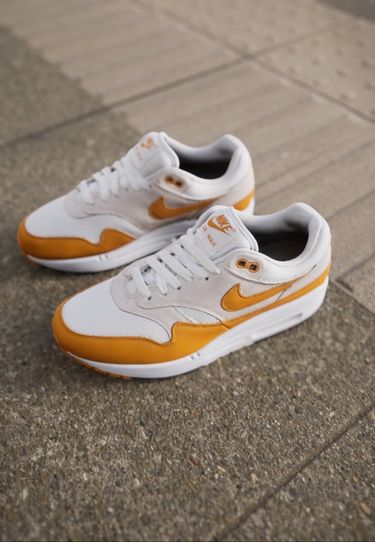 Nike Air Max sneakers hebben een wit en oranje kleurenschema, met textuur suède en mesh materialen, voorzien van veters en een zichtbare luchtkussenunit.
