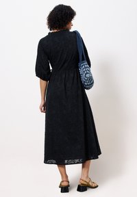 Schwarzes Midi-Kleid mit strukturiertem Blumenmuster, gepufften Ärmeln und tailliertem Schnitt. Akzentuiert mit einer blauen gewebten Handtasche und grünen Riemchensandalen.