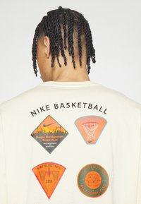 Krémová bavlněná mikina s kapucí, která má čtyři barevné nášivky s tématem basketbalu a značkový nápis "Nike Basketball" tučným černým písmem.
