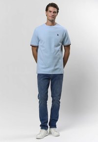 Ljusblå kortärmad T-shirt med rund halsringning, med en liten logotyp och randiga detaljer vid ärmarna, kombinerad med blå jeans och vita sneakers.