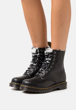 Musta nahkaiset nilkkurit, joissa on tekoturkiskaulus, pyöreä varvas, nauhakiinnitys ja keltaiset tikkauslangat pohjassa. Kestävä kumipohja tarjoamaan pitoa.