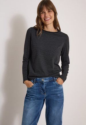 Femme souriante les mains dans les poches, portant un t-shirt à manches longues rayé noir et gris et un jean bleu denim sur un fond clair.