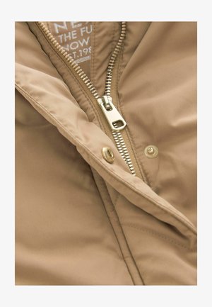 Lichtbeige jas met een gladde textuur, voorzien van een gouden rits en een label met witte letters dat gedeeltelijk zichtbaar is aan de binnenkant.