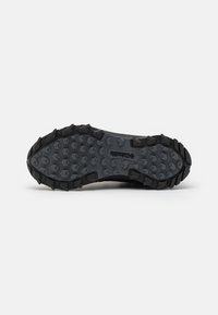 Semelle de chaussure noire robuste avec des crampons profonds hexagonaux et angulaires conçus pour la traction en extérieur, avec un logo de marque visible au centre.