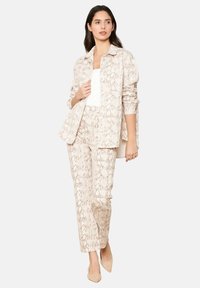 Costume en imprimé serpent beige avec une veste boutonnée et un pantalon fuselé, en tissu texturé, associé à un haut blanc et des talons nus.