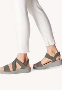 Sandalias verde oliva con tiras elásticas en cruz, diseño de punta abierta y suela gruesa gris. Combinadas con unos jeans blancos recortados.