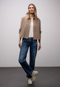 Femme debout, jambes croisées, portant une veste beige boutonnée, une chemise blanche, un jean bleu et des baskets blanches sur un fond uni.