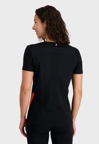 Black short-sleeve shirt with an orange accent on the side, curved panels on the back, and a small tri-color tag at the neckline.