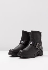 Bottines en cuir noir avec un bout arrondi, fermeture éclair sur le côté et une sangle brillante avec une boucle. Détails décoratifs avec des clous sur le bord supérieur.