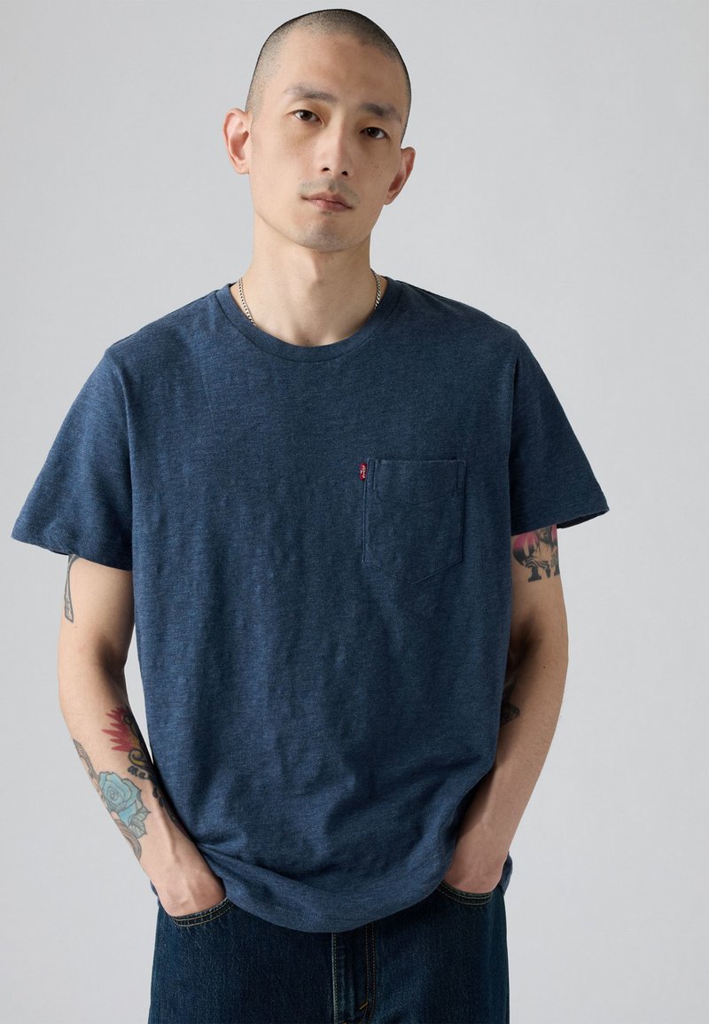 Mann mit Tätowierungen, trägt ein dunkelblaues T-Shirt mit Tasche und dunkle Jeans, steht mit den Händen in den Taschen vor einem schlichten Hintergrund.