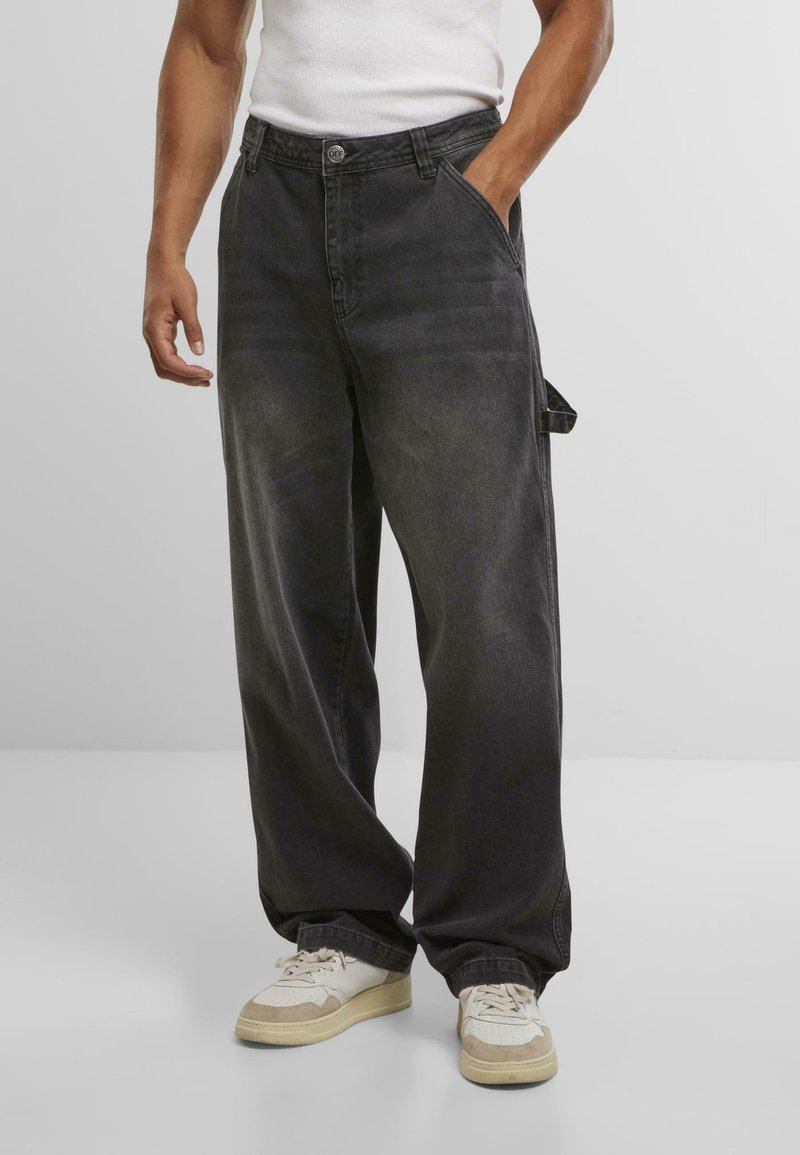 Losse, donkergrijze denim broek met knoopsluiting, voorzakken en een zijschoen detail, gecombineerd met witte en beige sneakers.