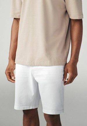 Korta vita denimshorts med rak skärning, i kombination med en beige kortärmad skjorta. Tyget verkar mjukt och avslappnat.