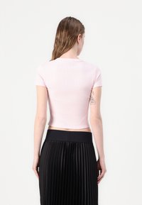 Kvinde med vådt hår iført en lyserød cropped top og sort plisseret nederdel, vendt væk, med en arm tatovering på hvid baggrund.