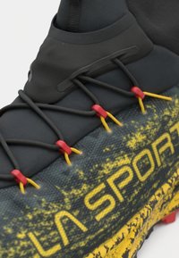 Prim-plan al unei încălțări sport negre și galbene, cu sireturi negre trecute prin găurelele roșii și galbene, pe o parte superioară din material texturat.