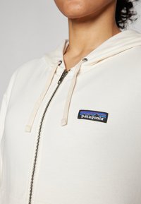 Kremasti zip-up dukserica s vezicama, sa crnim Patagonia logom i dizajnom planine u bojama. Glatka tkanina, ležeran kroj.