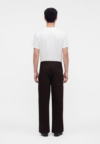 Homme debout, de dos, portant un t-shirt blanc uni à manches courtes et un pantalon noir ample avec des chaussures noires sur fond blanc.