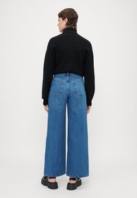 Pulover texturat negru, cu guler înalt, asortat cu blugi largi din denim albastru. Blugii au buzunare pe spate și margini tivite. Pantofi negri cu platformă.