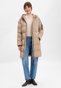 Beige gepolsterter Mantel mit Kapuze, der mit einem leichten Pullover und blauen Jeans getragen wird. Er hat Reißverschlusstaschen und schwarze Akzente. Glatte Textur.