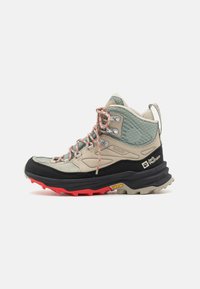 Hochschuh-Wanderschuh mit beigem Obermaterial und strukturierten grünen Akzenten, schwarzer und roter Sohle sowie leuchtend roten Schnürsenkeln. Vibram-Außensohle für Grip.