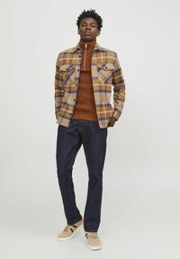 Bruine geruite flanellen shirt met twee borstzakken over een roestkleurige coltrui, gecombineerd met donkere spijkerbroek en beige sneakers met maroon accenten.