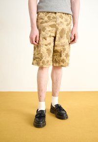 Beige camouflage cargo shorts with side pockets and a textured fabric, paired with black shoes and white socks. Flooring is yellow.