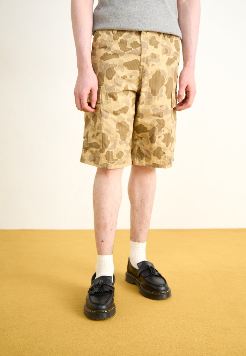 Beige camouflage cargo shorts with side pockets and a textured fabric, paired with black shoes and white socks. Flooring is yellow.