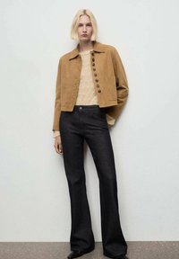 Jachetă din suede maro cu nasturi mari, purtată cu un pulover tricotat crem și blugi negri evazați. Design simplu cu linii curate.