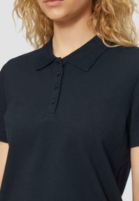Blonde persoon met een zwart kort-om-mouw polo shirt met vier knopen en een klassieke kraag, zichtbaar van nek tot taille.