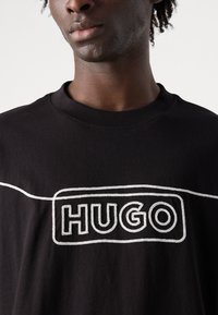 Juodas marškinėlis su apykakle turi išskirtinį baltą išsiuvinėtą "HUGO" logotipą ir baltą liniją per krūtinę. Minkšta medvilnės medžiaga.