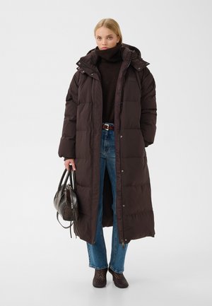 Femme portant un long manteau matelassé foncé, un col roulé noir, un jean bleu et des bottines, tenant un sac à main foncé texturé.
