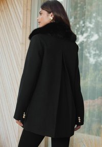 Čierny kabát s umelým kožušinovým golierom, so štruktúrovaným dizajnom, jedným sklonom vzadu a pozlátenými gombíkovými akcentmi na rukávoch. Hladká textúra.