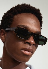 Lunettes de soleil en écaille de tortue avec des montures noires angulaires, des verres sombres et des accents texturés. Vue en gros plan axée sur le design des lunettes.