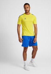 Camiseta atlética amarilla de manga corta, pantalones cortos azules con rayas laterales y zapatillas blancas. Diseño simple y funcional para ropa deportiva.